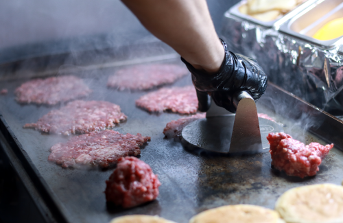 A fry chef wearing black nitrate gloves using a cast iron press to flatten smash burgers on a griddle.