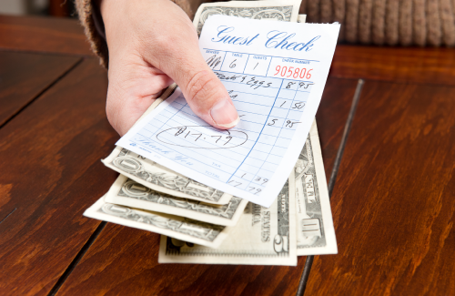 A hand holding a collection of USA dollar bills and a guest check, implying they are paying their bill.