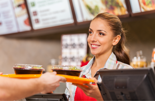 A female fast food worker standing behind the register and passing an orange tray with two meals on it to a man off screen. She is smiling.