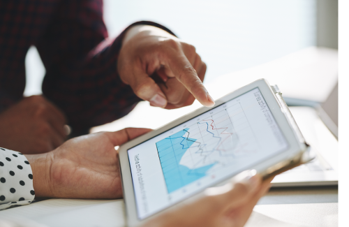 a woman holding an ipad with a line graph displayed on it. A man is sitting next to her and pointing at the screen to indicate data change.