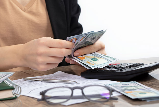 A close-up image of a person counting cash while using a calculator on a wooden desk. The scene includes eyeglasses, documents, and a pen in the background, emphasizing a workspace setting focused on financial calculations or cash management.